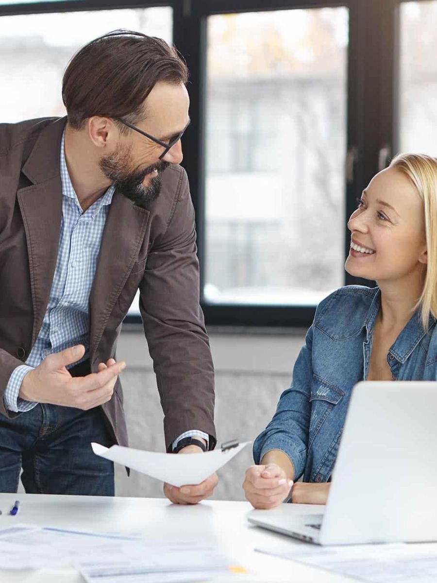 Handsome mature male financier and his female secretary work with documents, discuss something actively, have happy expressions, use modern laptop and internet for surfing Internet. Career concept